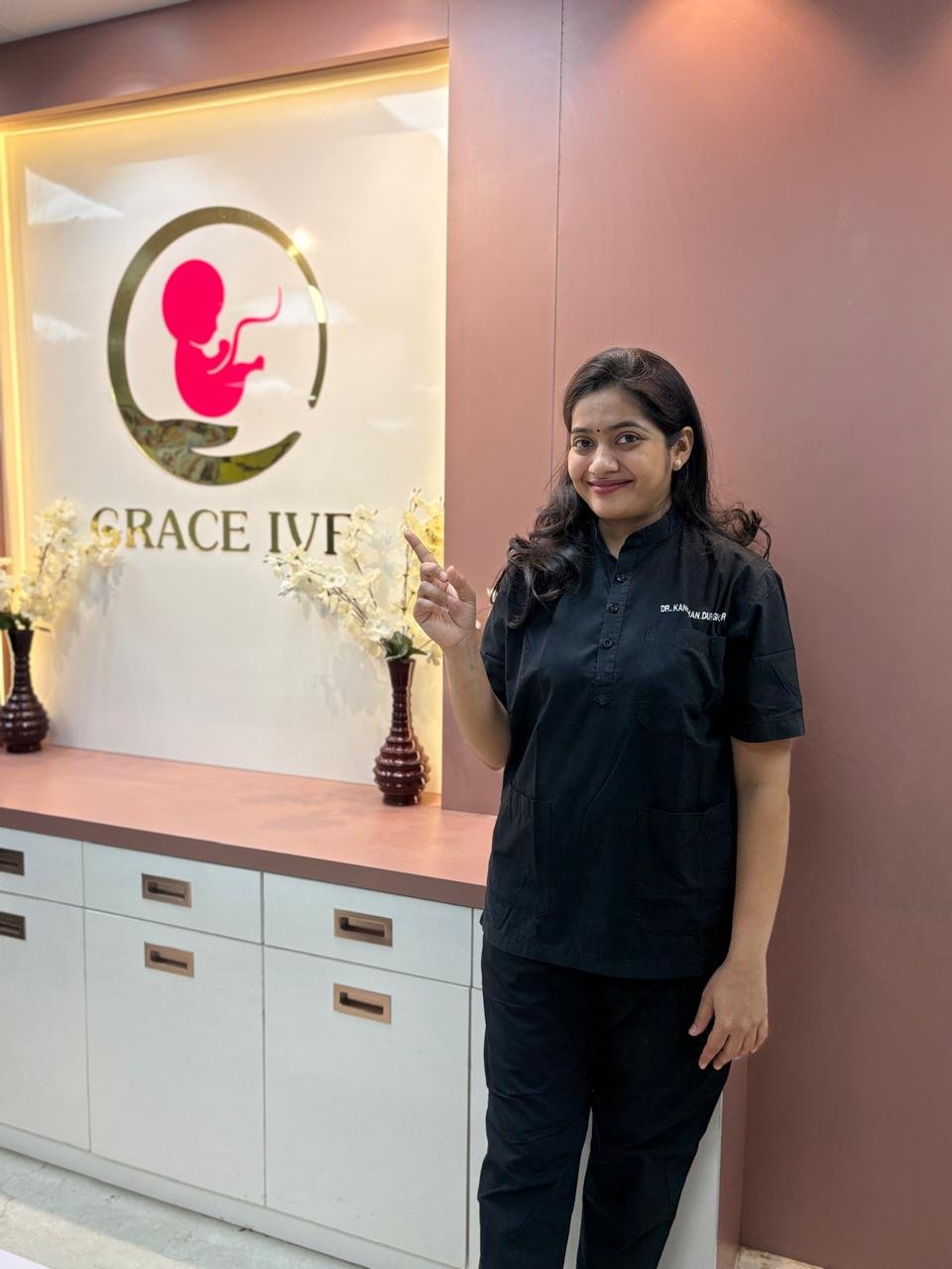 A female doctor in black medical scrubs standing inside Grace IVF Clinic, smiling and pointing upward while posing beside the clinic logo.